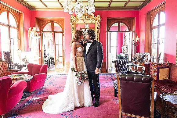 Wedding kiss portrait of bride and groom kiss beside a gilded mirror, her rose bouquet in hand under a crystal chandelier in a pink lounge