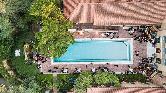 Poolside wedding reception with round tables and white tablecloths by a swimming pool, string lights glowing in a courtyard with trees and hedges