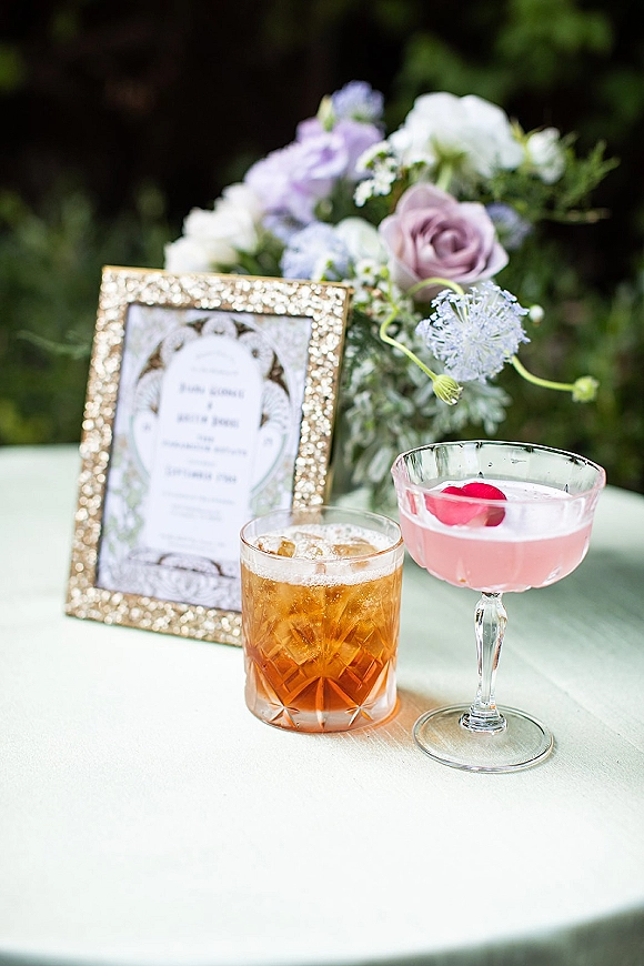 Signature wedding cocktails beside a gold-framed signature drink menu, featuring a pink coupe and whiskey rocks glass with fruit garnish on white linen