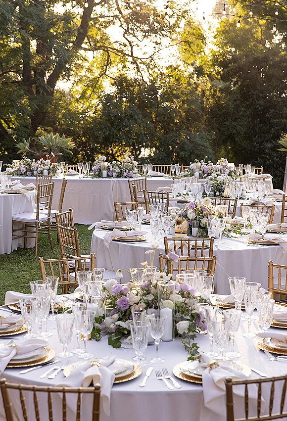 Reception tablescape with white linens, gold chiavari chairs, floral garlands and taper candles on a garden lawn under string lights at sunset