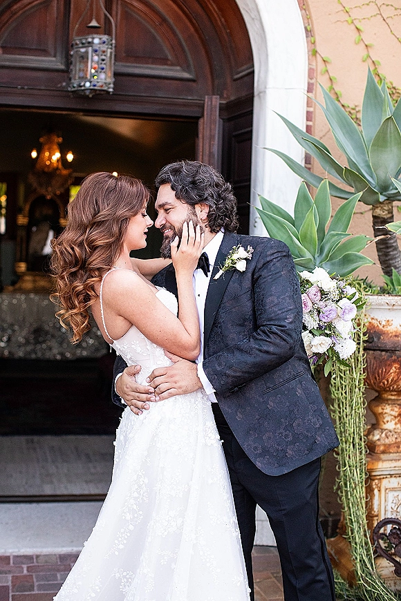 Couple portrait of bride and groom embrace with a forehead touch, bride in lace gown holding bouquet by an arched doorway with vines