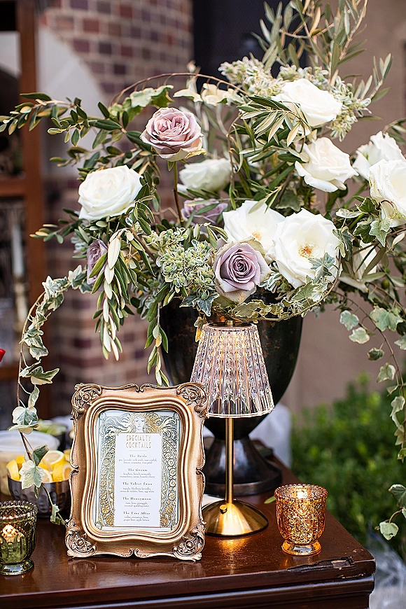 Cocktail table decor with a wedding cocktail menu sign in a gold frame, roses and ivy urn centerpiece, candles, and lemon slices by a brick wall