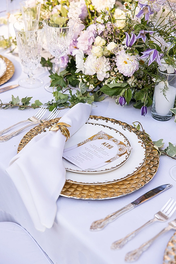 Wedding tablescape with gold and white tablescape details, lavender and white floral centerpiece, crystal goblets, and menu card in outdoor light