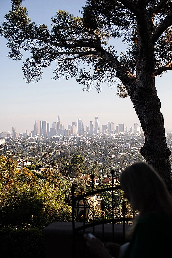 City skyline view from a wedding venue city view terrace, framed by a large tree and hillside neighborhood, with an outdoor lantern on the patio railing