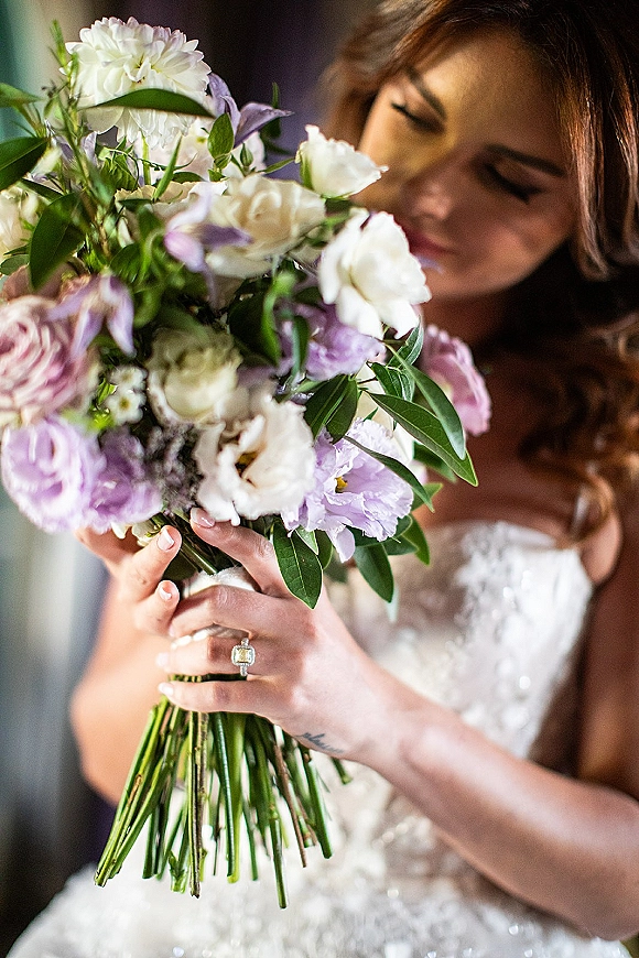 Bridal bouquet of white and lavender bouquet blooms with greenery, held against a sparkly gown bodice and engagement ring in window light