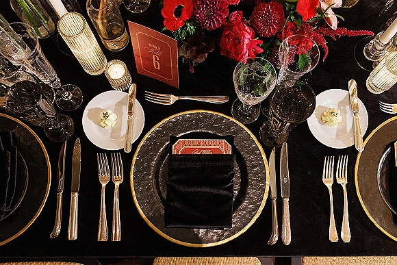 Reception tablescape with a black and gold wedding table setting, crystal glasses, taper candles, and a red floral centerpiece with place cards