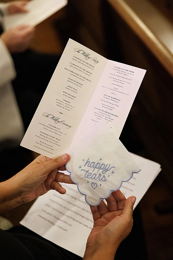 Wedding ceremony program resting on a happy tears handkerchief with embroidered text, in church pews during an indoor ceremony