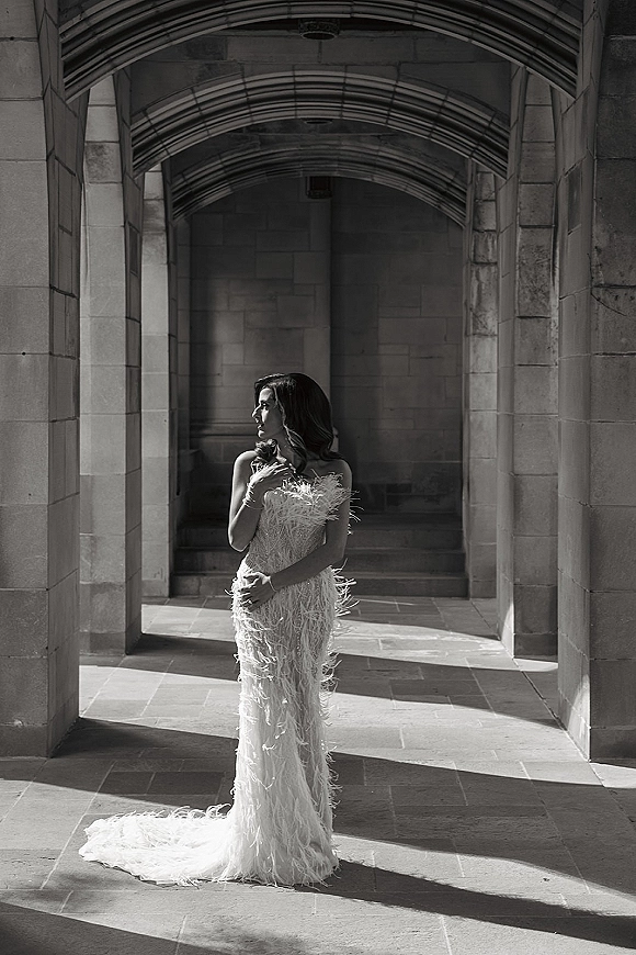 Bridal portrait of a bride in a strapless feather wedding dress with long train, standing in a stone arch colonnade with shadows