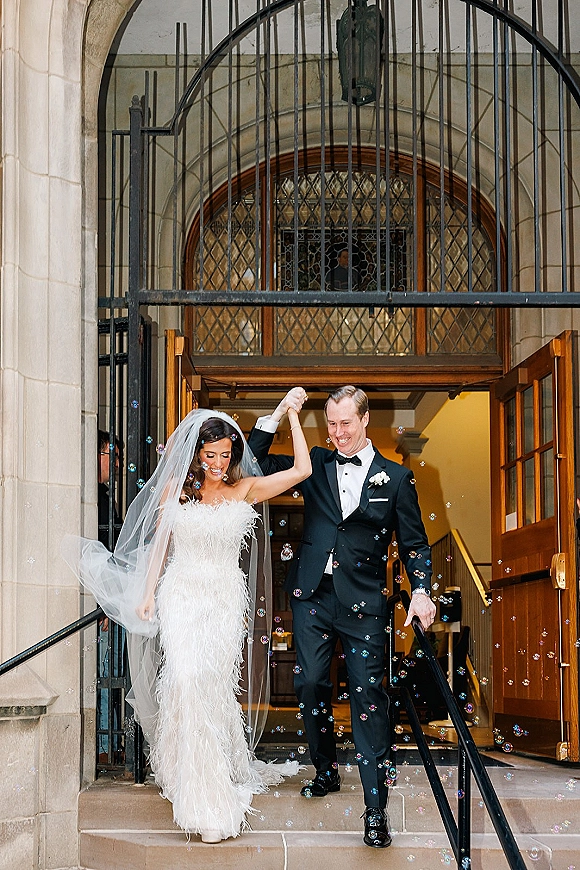 Wedding recessional as newlyweds walking out hand in hand, bride in veil and strapless gown, through stone church entrance amid bubbles