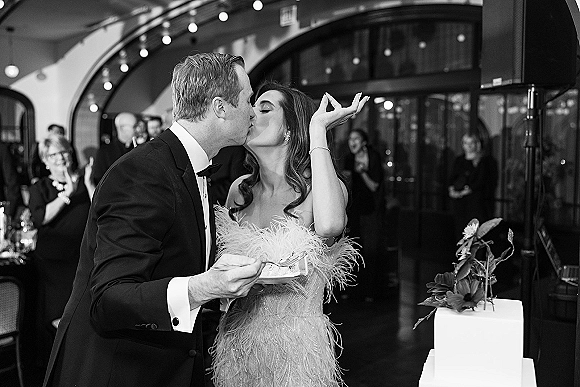 Wedding cake cutting as bride in feather dress and groom in tuxedo share a kiss by floral-topped cake under string lights indoors