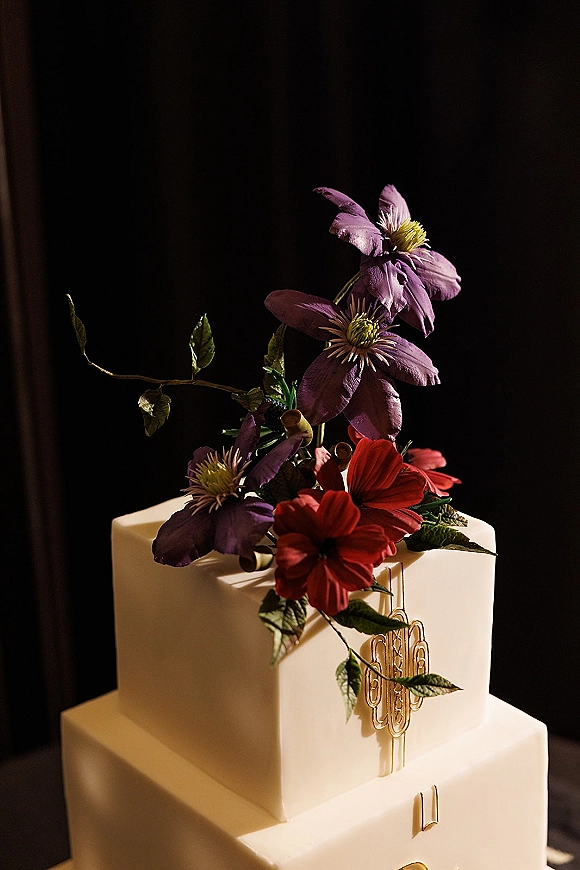 Wedding cake with a floral cake topper and gold monogram, white fondant tiers with sugar flowers against a dark moody backdrop