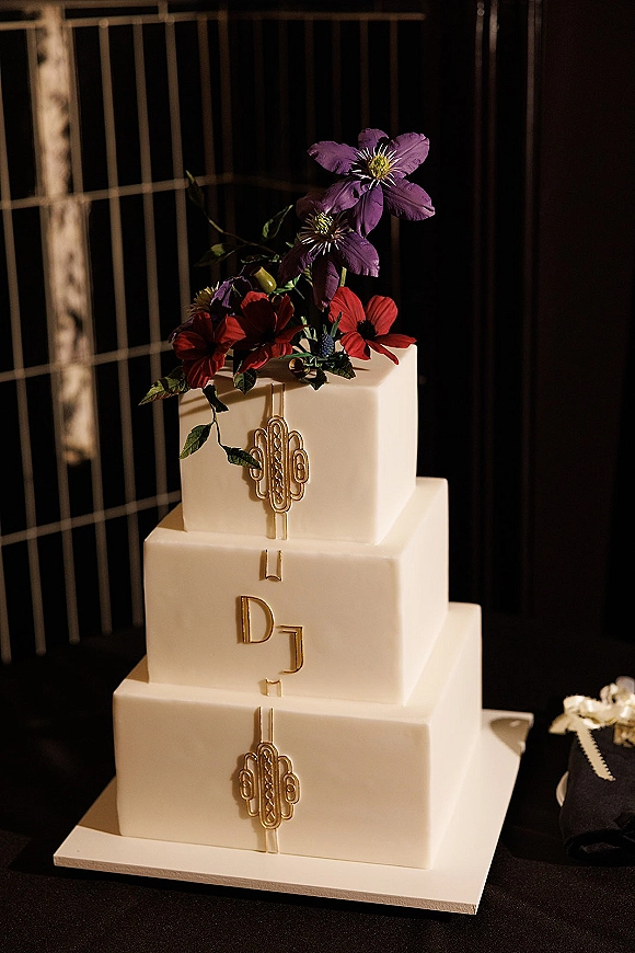 Wedding cake with white fondant in three square tiers, gold monogram and geometric piping, topped with purple and red flowers on a black tablecloth