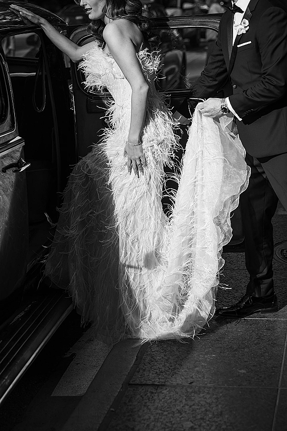 Wedding exit moment as groom helps bride into a vintage wedding car, her long veil and feather strapless dress by the open door curbside