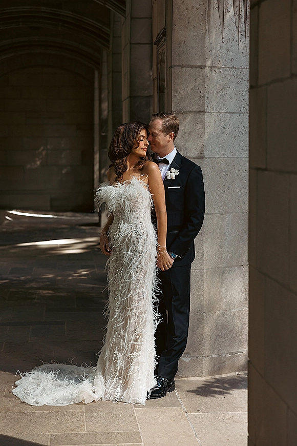 Couple portrait of groom kissing bride’s forehead as she leans on him in a feather wedding dress under stone columns and dappled light