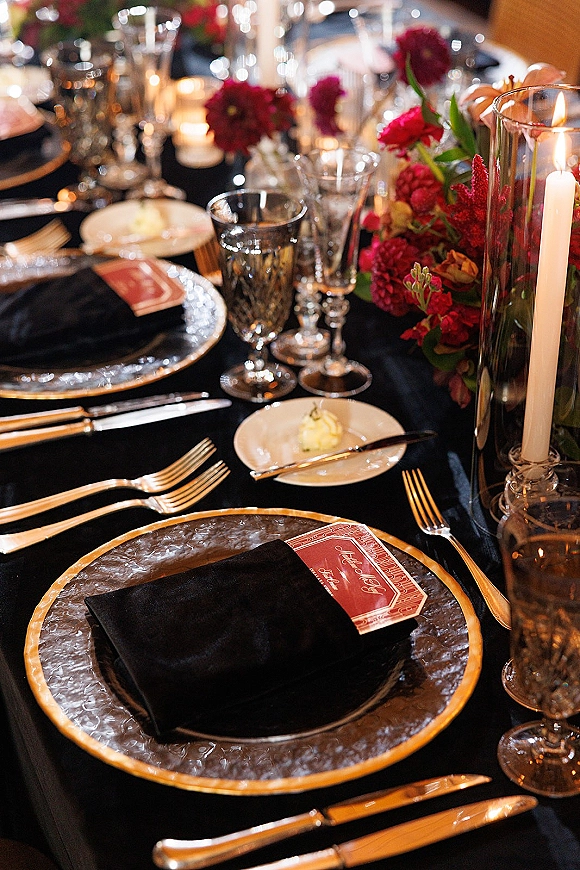 Reception tablescape with black and gold wedding table details, patterned plates, gold flatware, red flowers, and candlelit bokeh backdrop
