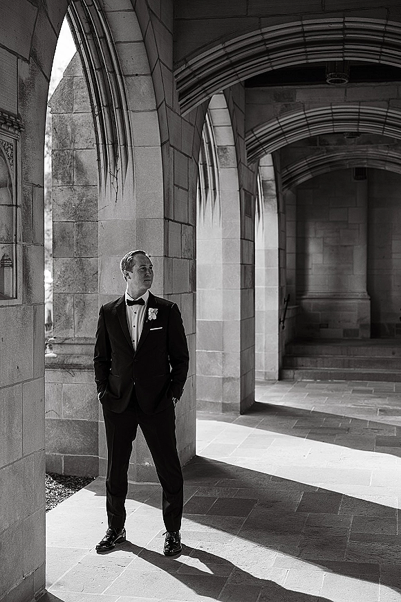 Groom portrait of a black tie groom in a tuxedo with white rose boutonniere, leaning in a sunlit stone archway hallway with shadows