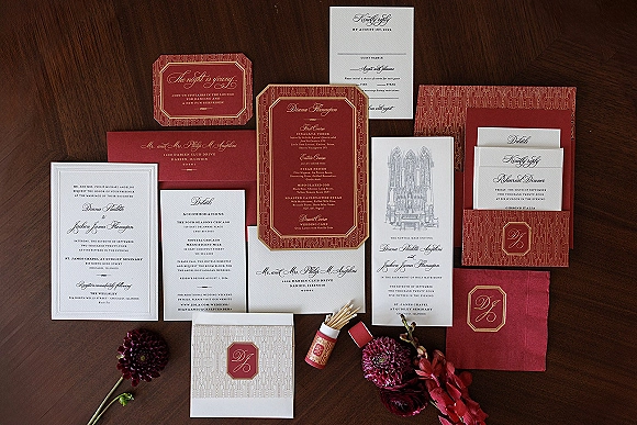 Invitation suite with burgundy wedding invitations, envelopes and reply card, monogram seal, matchbook, and flowers on a dark wood table