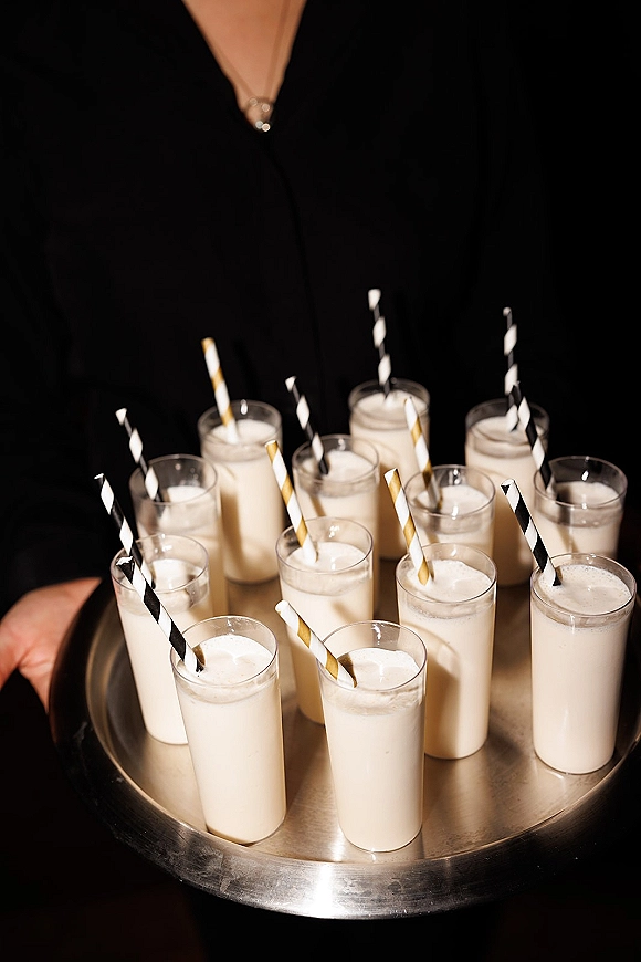 Wedding drink tray of wedding welcome shots in shot glasses with creamy drinks and striped paper straws held against a dark background