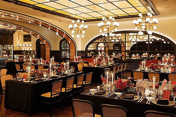 Reception tablescape with black wedding table linens, red-pink florals, taper candles and glass vases under chandeliers in an indoor hall