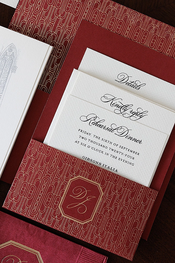 Invitation suite with burgundy wedding invitations and gold foil monogram, calligraphy cards, and patterned envelopes on dark wood surface