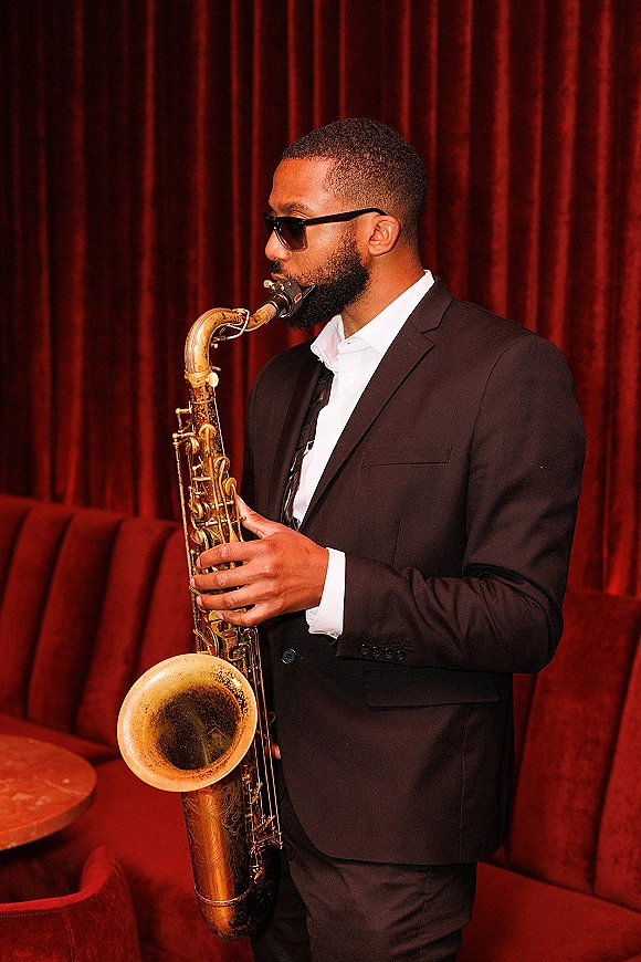 Wedding saxophonist playing during wedding cocktail hour music in a black suit and sunglasses against red velvet curtains and banquette seating
