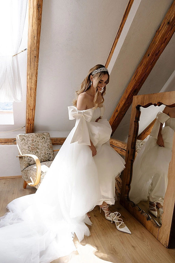 Bridal portrait of a bride in a strapless wedding dress with oversized bow sleeves, standing by a floor mirror in a sunlit attic room