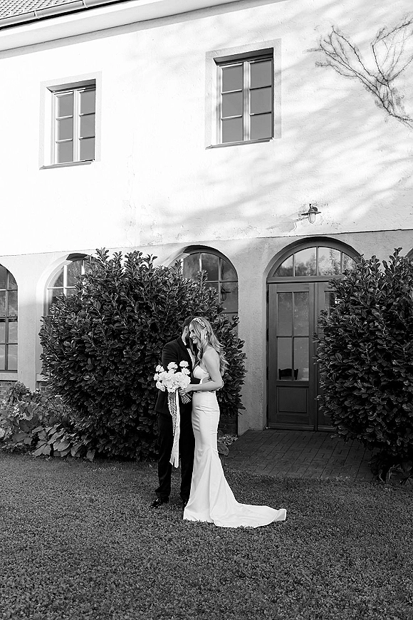Couple portrait of bride and groom in an embrace with a forehead kiss, bride holding bouquet by a stucco building with arched doorway