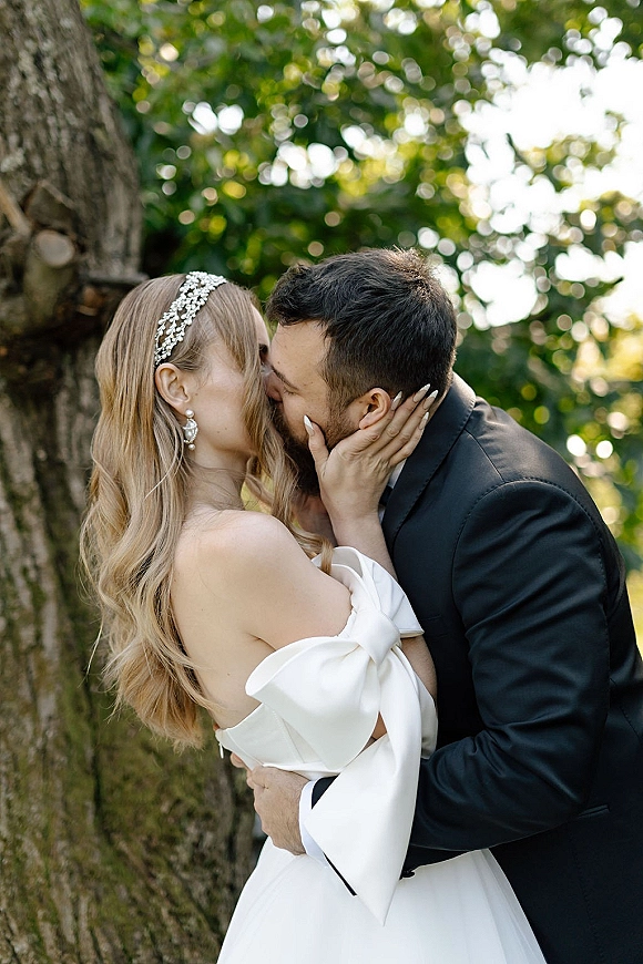 Wedding kiss portrait of bride and groom kissing, bride in strapless dress with bow sleeve and crystal headband in sunlit garden bokeh