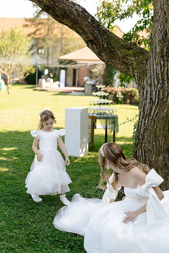 Flower girl moment as the bride kneels beside her on a garden lawn, veil trailing, with a champagne tower table in the background