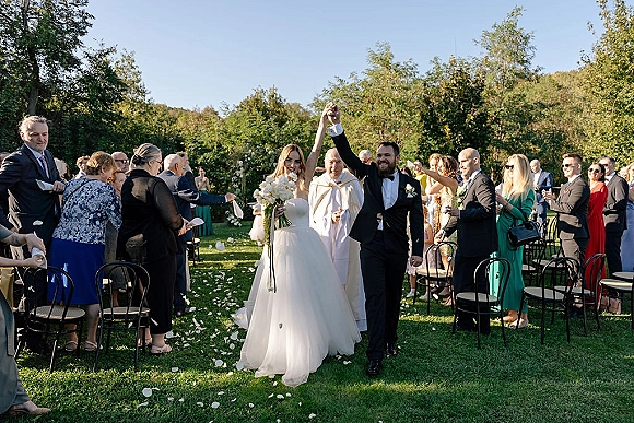 Wedding recessional as bride and groom walk the aisle with raised hands, she holds a bouquet, guests cheer on a sunny lawn under blue sky