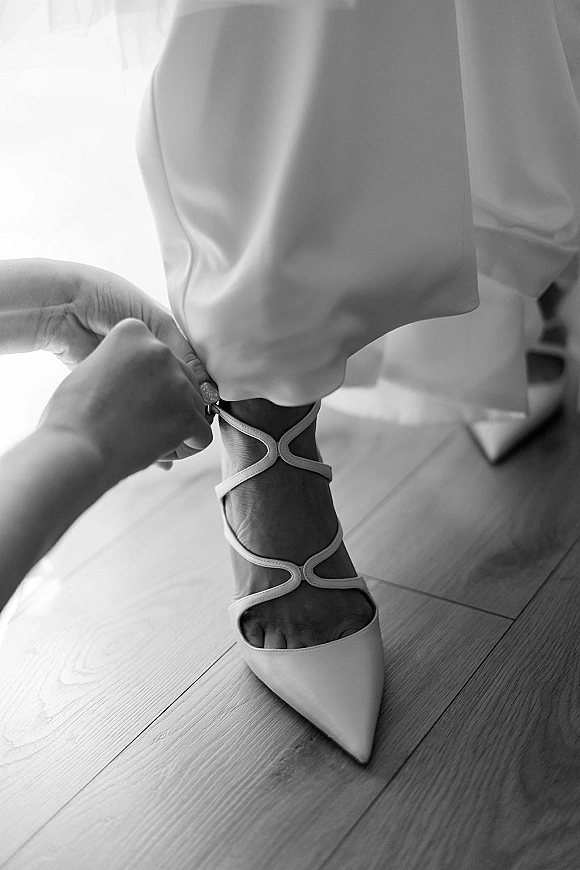 Bridal shoes in pointed toe heels as hands fasten ankle straps over bridal pants on a wood floor in soft window light