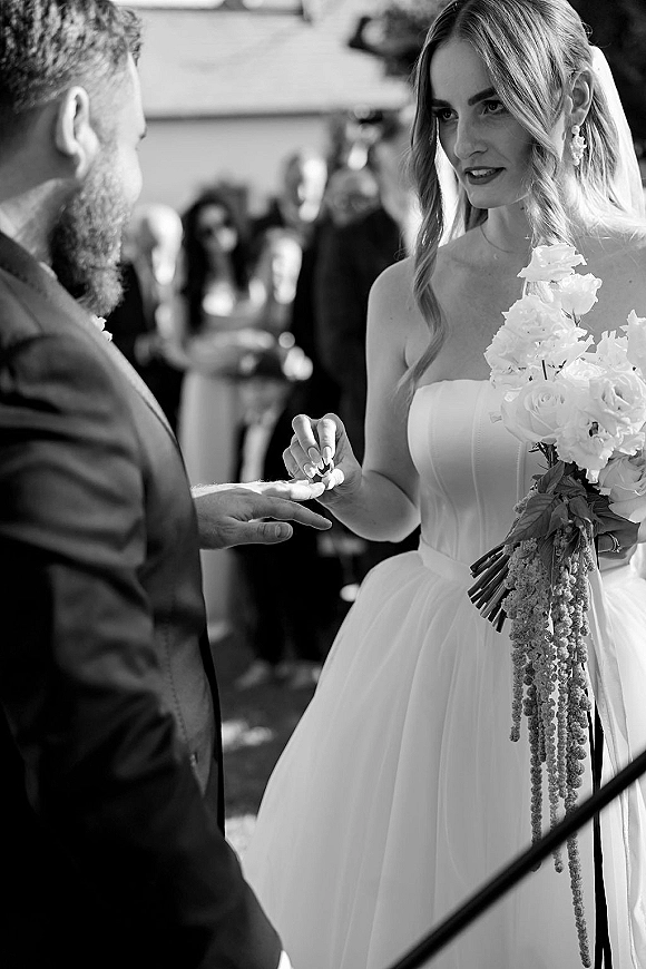 Ring exchange as the bride slips a wedding ring exchange onto the groom’s hand, holding a white bouquet in an indoor ceremony with guests blurred
