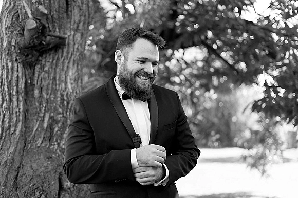 Groom portrait in a black tuxedo, smiling with boutonniere and bow tie while leaning against a tree amid lush greenery outdoors
