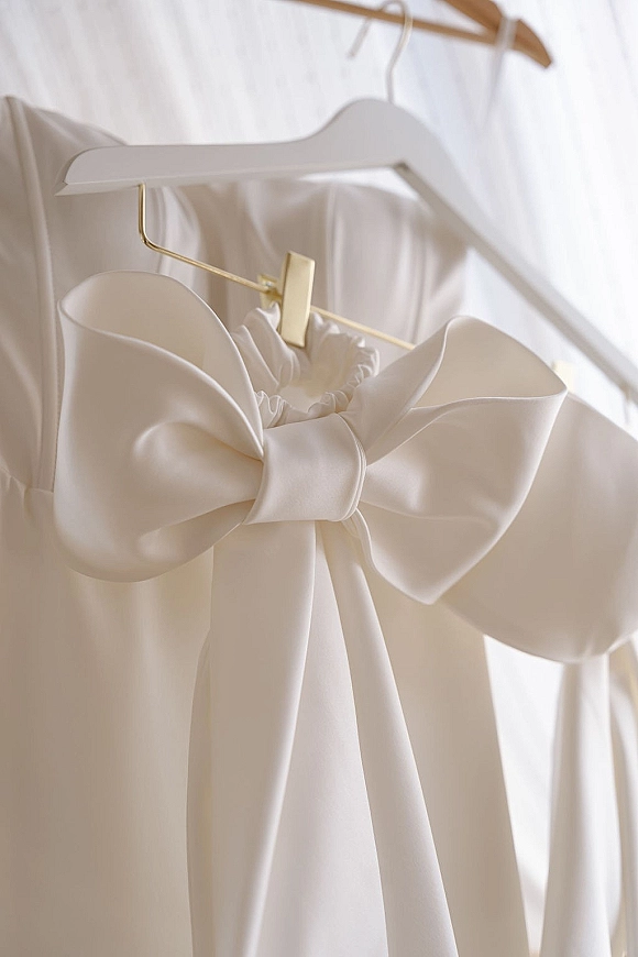 Wedding dress close-up with a satin bow wedding dress detail, fabric buttons and gold hanger hook, hanging against a white wall with garments