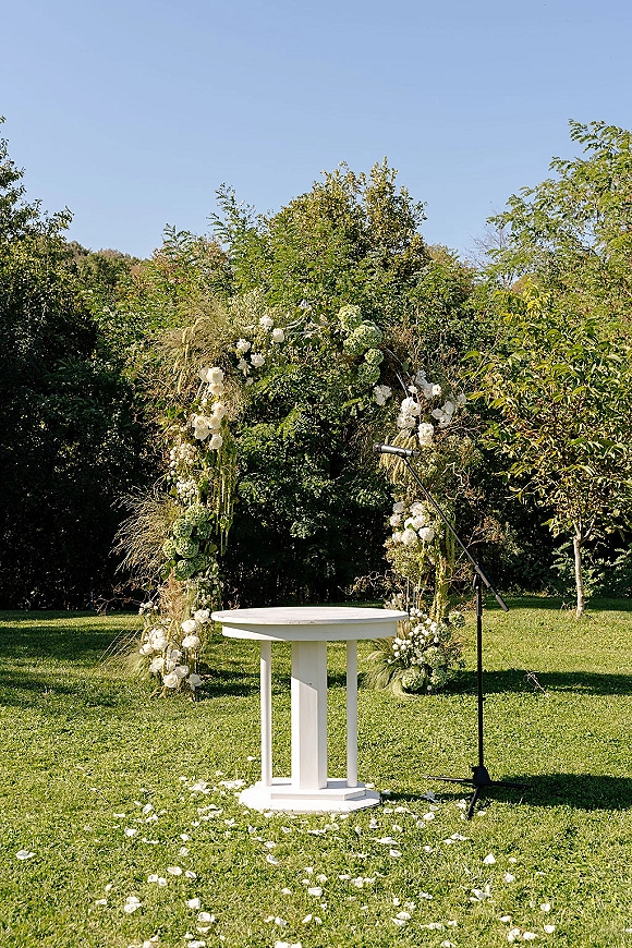 Ceremony altar decor with a white floral arch and greenery, microphone stand, and petal-strewn aisle on a sunny lawn under blue sky