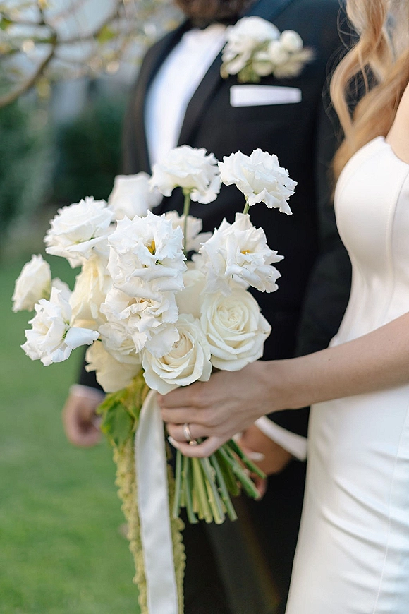 Bridal bouquet of white rose bouquet and lisianthus with trailing greenery, held by bride in satin dress beside groom on a lawn
