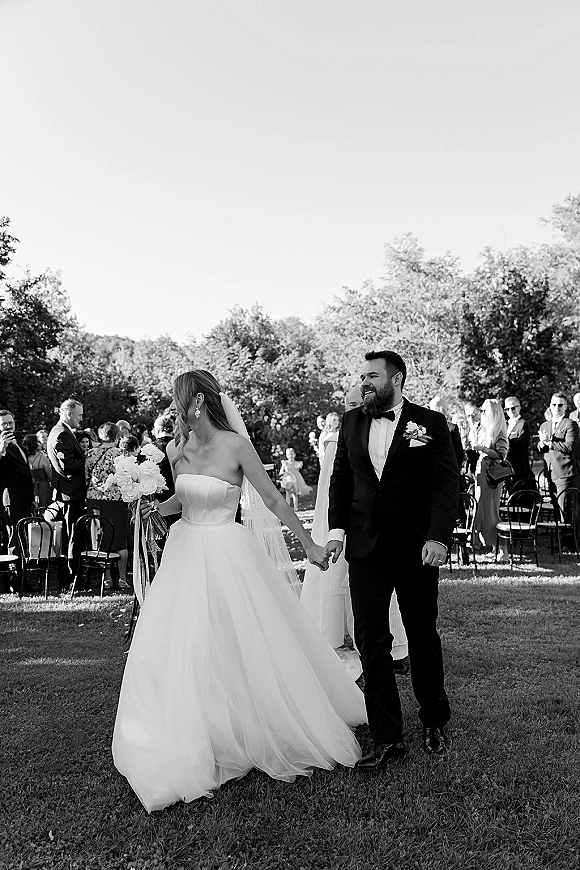 Wedding recessional as bride and groom walk the aisle holding hands, bride in strapless gown and cathedral veil with bouquet, guests on lawn