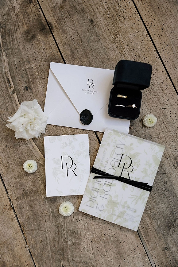 Invitation suite wedding invitation flat lay with envelopes, monogram wax seal, ribbon, white flowers, and rings in a velvet box on rustic wood floorboards