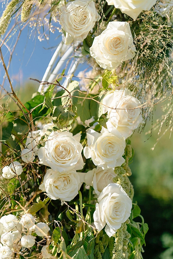 Wedding arch flowers with white rose clusters and greenery vines on an arch frame, set outdoors against blue sky and distant greenery