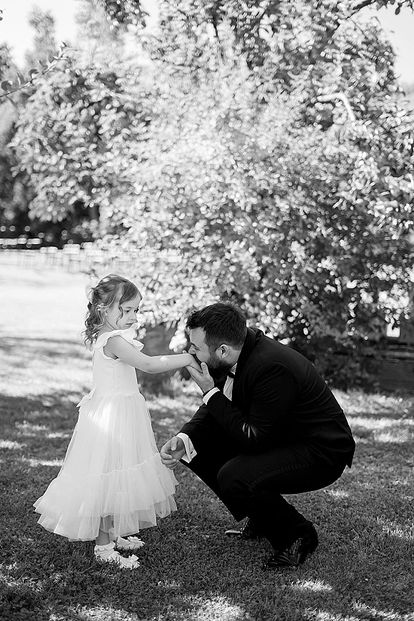 Wedding family moment as groom and flower girl share a sweet hand kiss on a garden lawn, his black tuxedo and her tulle dress with a hair bow