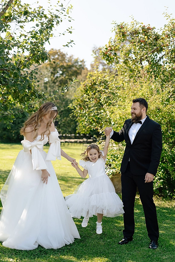 Wedding family portrait of bride and groom and child holding hands as the flower girl jumps on a sunlit garden lawn with trees