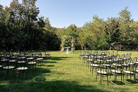 Outdoor ceremony setup with backyard wedding ceremony chairs lining a grass aisle to a floral arch, petals scattered on the lawn under blue sky