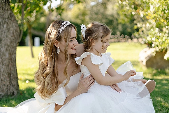 Bride with flower girl seated on her lap, both in white dresses with a pearl headband accent, laughing on a sunlit garden lawn
