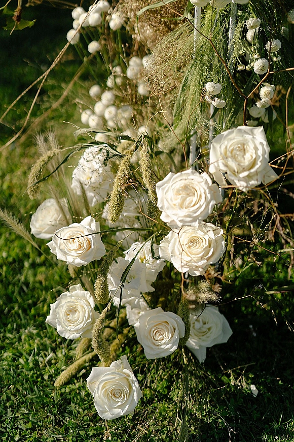 Wedding arch florals with white rose wedding arch arrangement of baby’s breath, dried grasses, and greenery on a metal frame over grass lawn