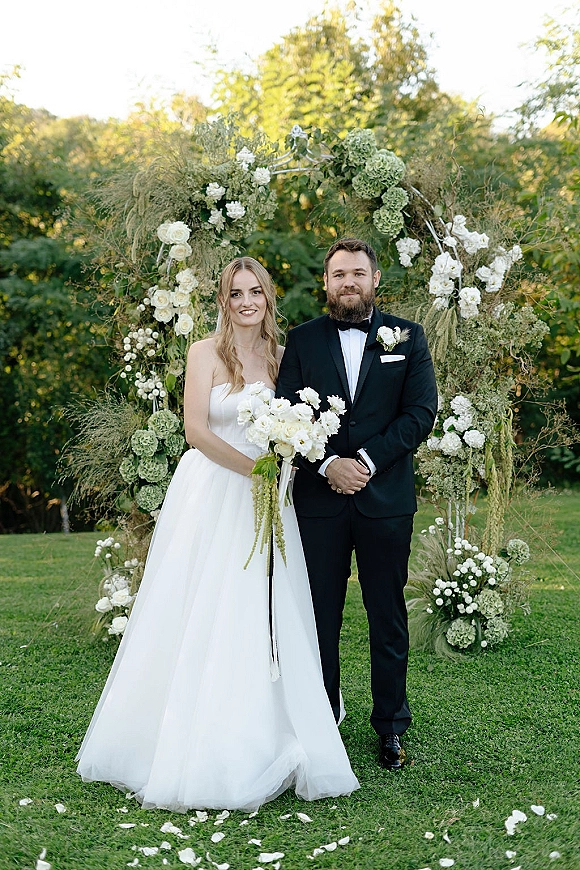 Couple portrait of bride in strapless gown holding a white rose bouquet beside groom in black tuxedo under a floral arch in garden daylight