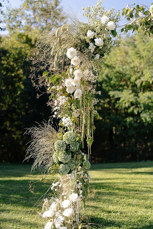 Wedding arch flowers on an asymmetrical wedding arch with white roses and greenery, cascading over a garden lawn with trees and open sky
