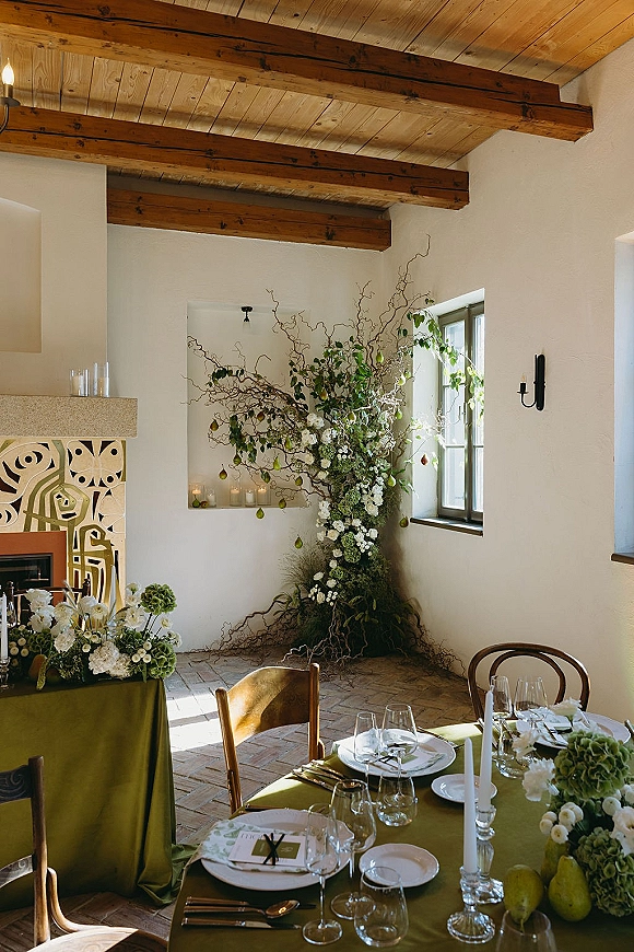 Reception tablescape with green wedding tablescape linens, pears, white florals, and taper candles on a long table beneath a wood-beam ceiling