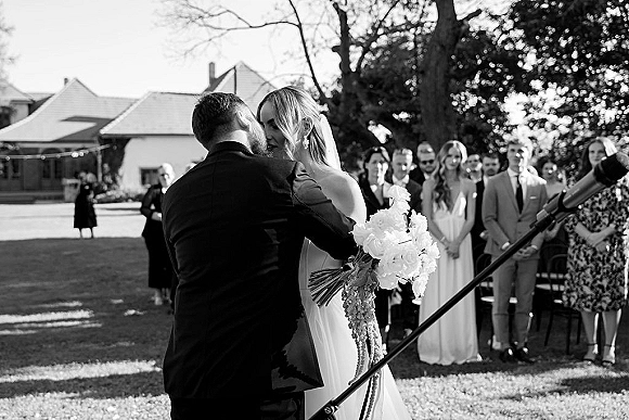 Ceremony kiss moment as bride and groom share their first kiss, her bouquet and veil visible, guests seated on a tree-lined lawn
