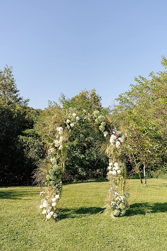 Wedding ceremony arch with white flowers and greenery, with pampas grass accents on a lawn under blue sky and trees