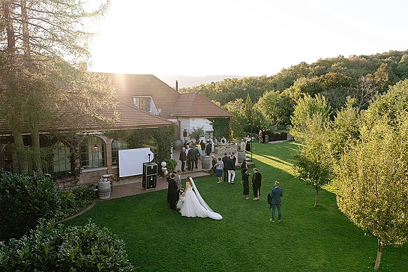 Outdoor wedding reception with wedding dress long train as guests mingle around cocktail tables on a sunlit lawn by trees and patio